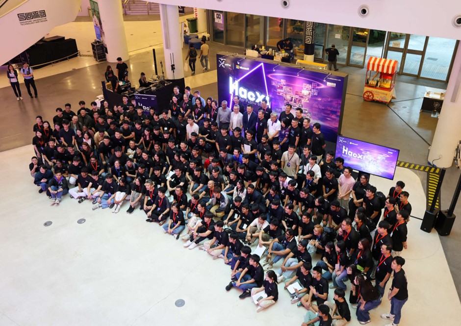 A large group of people pose for a group photo in an event hall with a large "HacX" sign in the background.