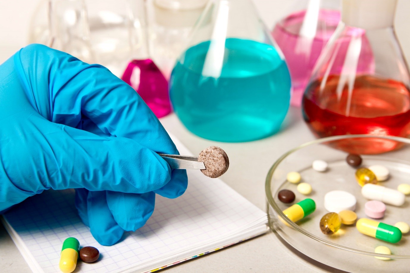 A gloved hand holds a pill with tweezers in a lab setting, with capsules, tablets, and flasks containing colourful liquids in the background.
