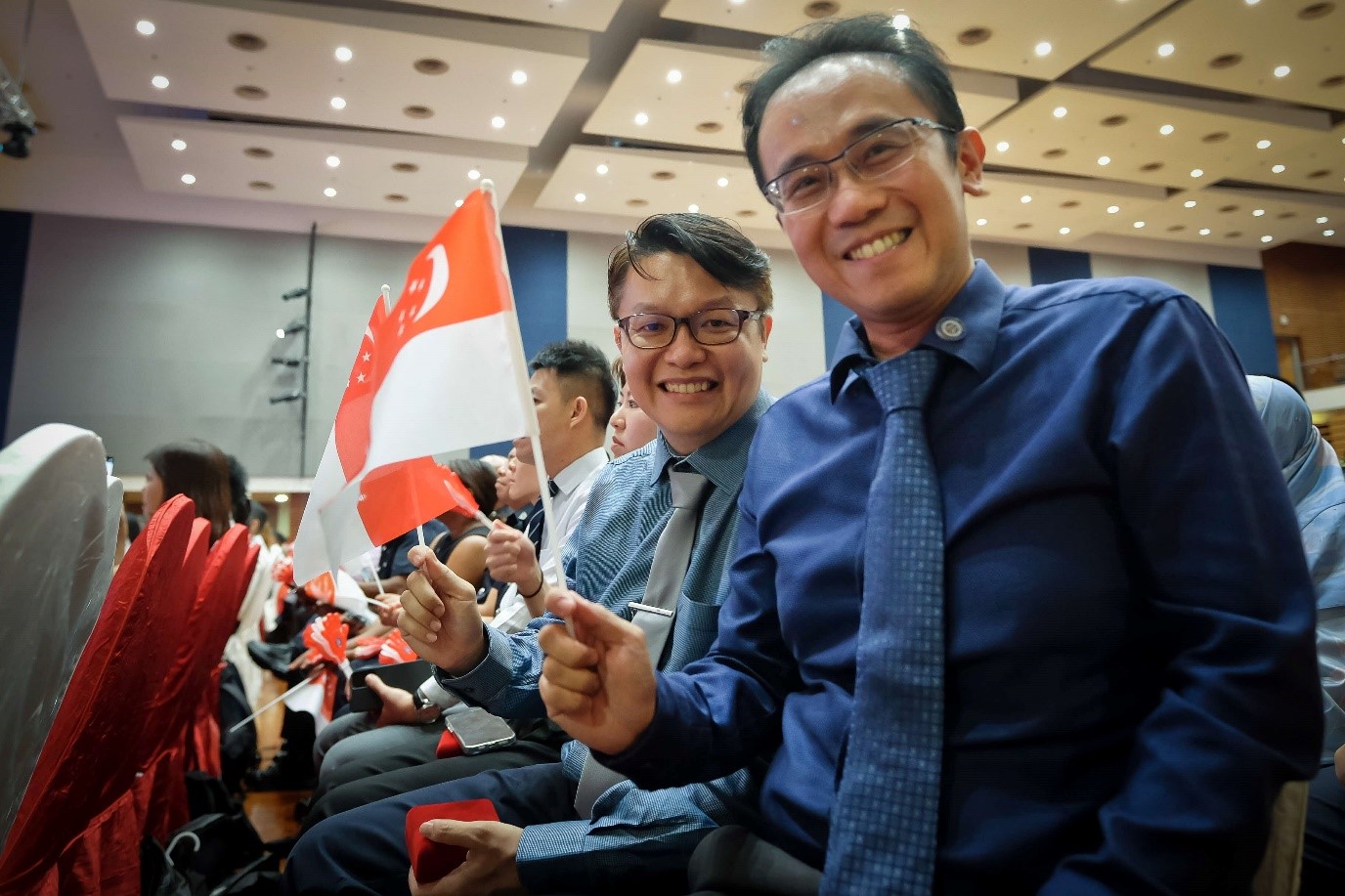 Two men sitting in a hall waving a mini Singapore flag.