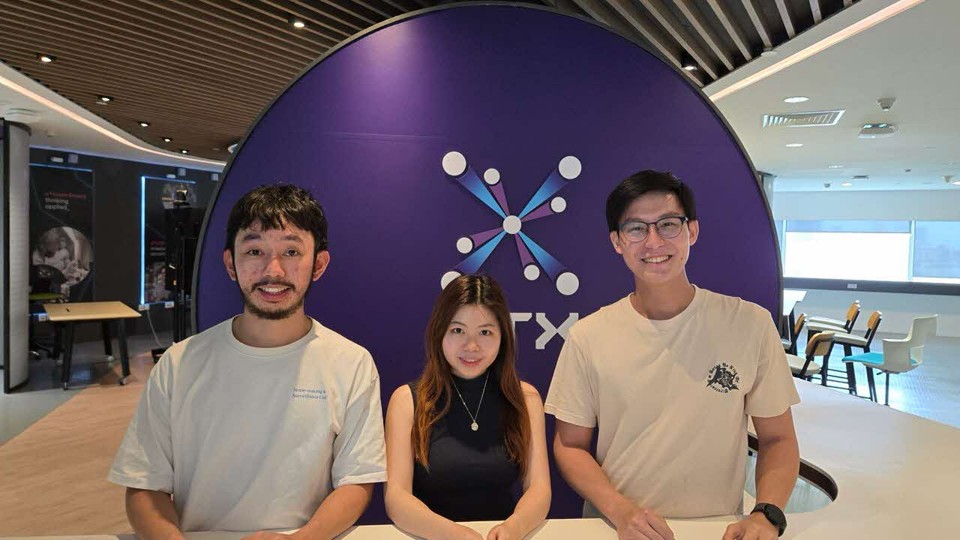 Three people, two men and one woman, stand in front of a large purple backdrop with the HTX logo.
