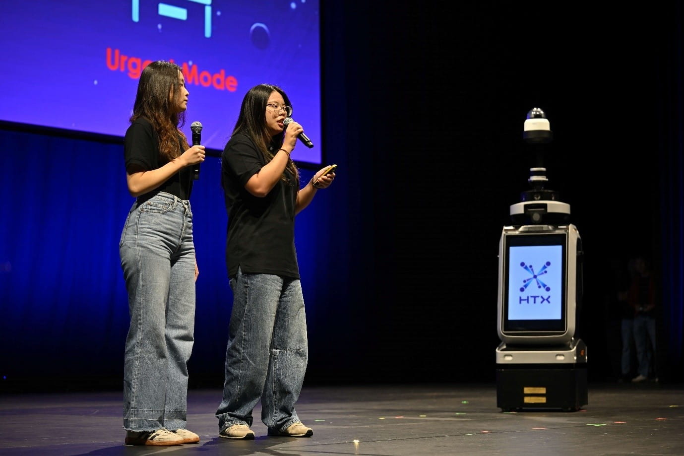 Two people on stage next to a robot displaying an "HTX" logo on its screen.