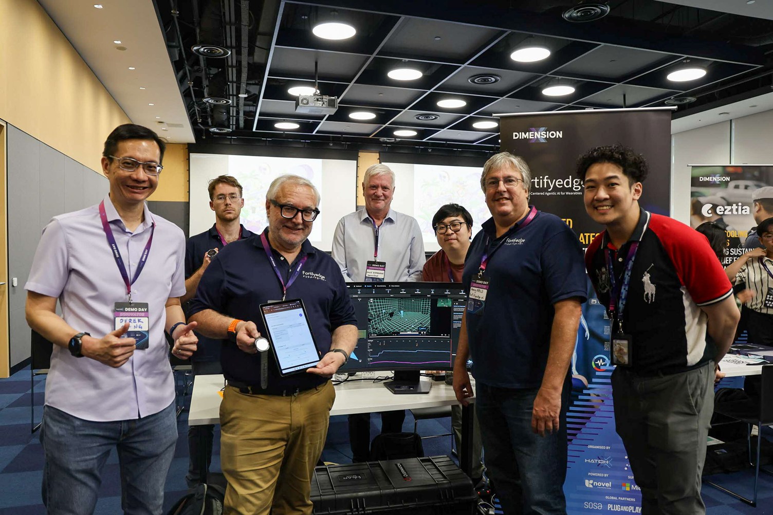 A group of men standing together and smiling in front of a display booth with one man holding a tablet.