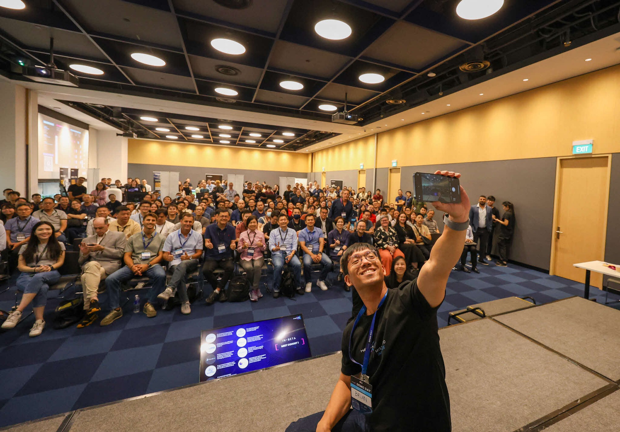 A man taking a selfie on stage with a large audience seated behind him.