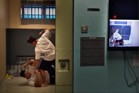 Two men inside a cell, shown on a CCTV monitor outside.