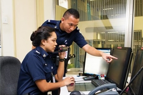 Two uniformed officers in a control room, with one speaking into a radio and the other pointing at a computer screen.