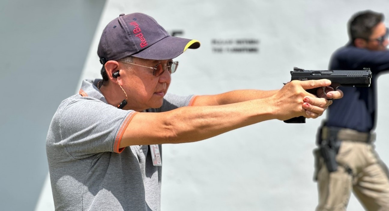 A man wearing a cap and sunglasses is seen in profile aiming a handgun at a firing range.