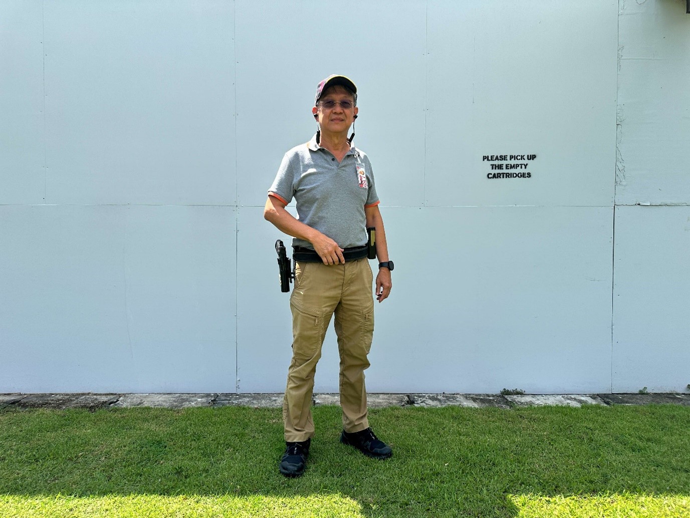 A man in a polo shirt and khaki pants with a holstered pistol stands in front of a white wall.