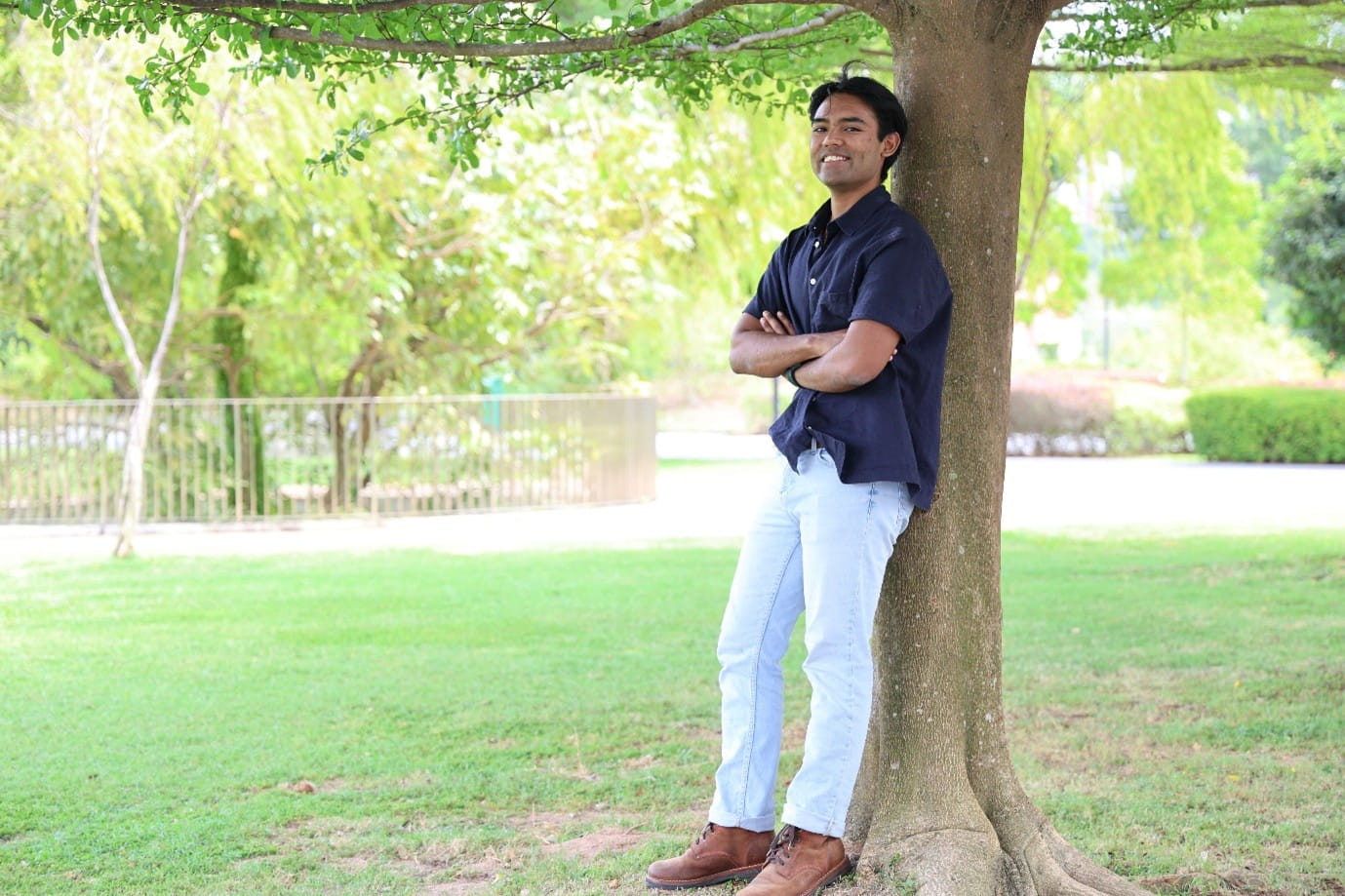 A young man in a navy shirt and jeans leans against a tree in a sunlit green park.
