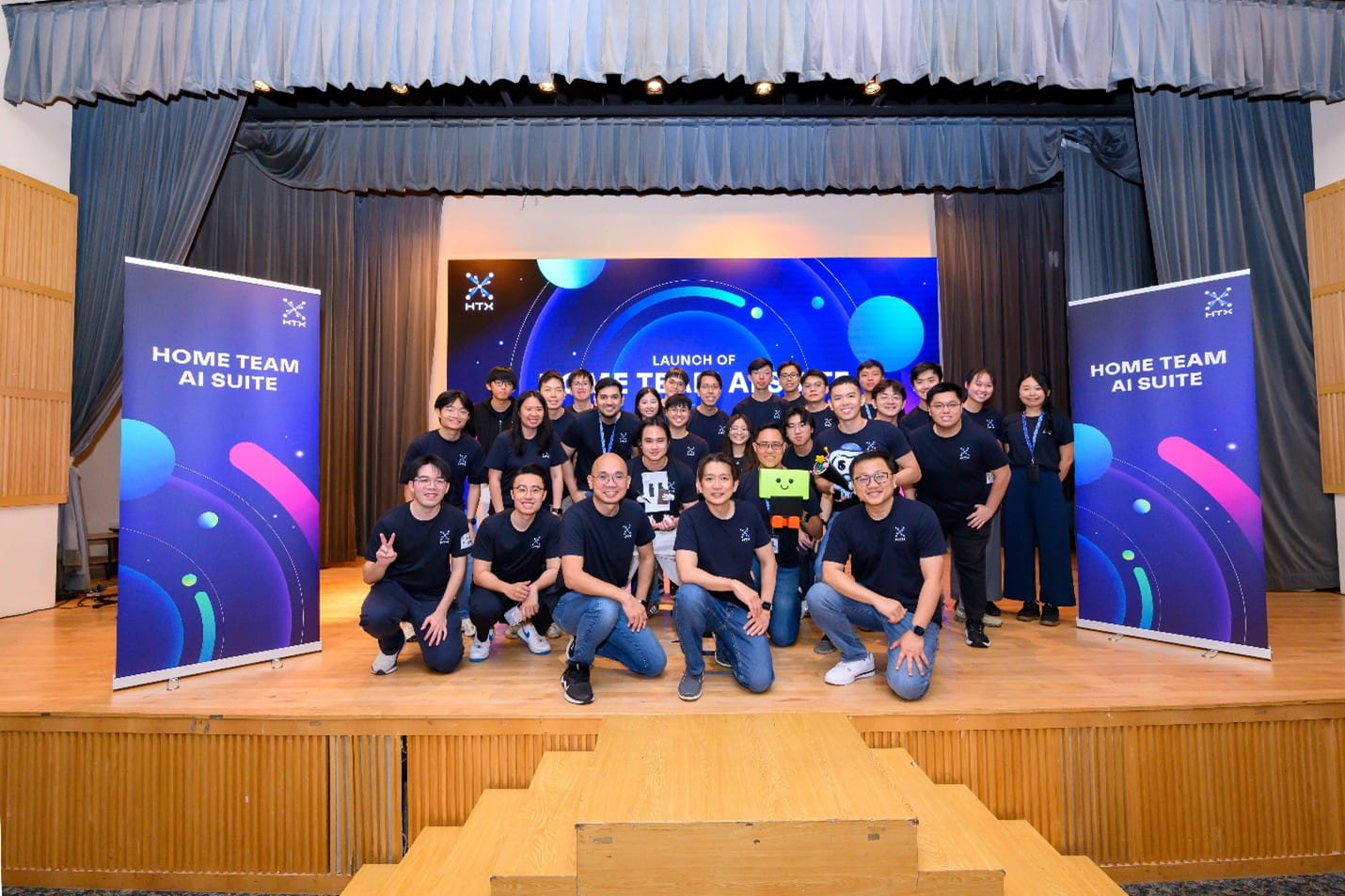 A large group of team members in matching dark HTX t-shirts posing for a photo on stage during the launch of Home Team AI Suite.