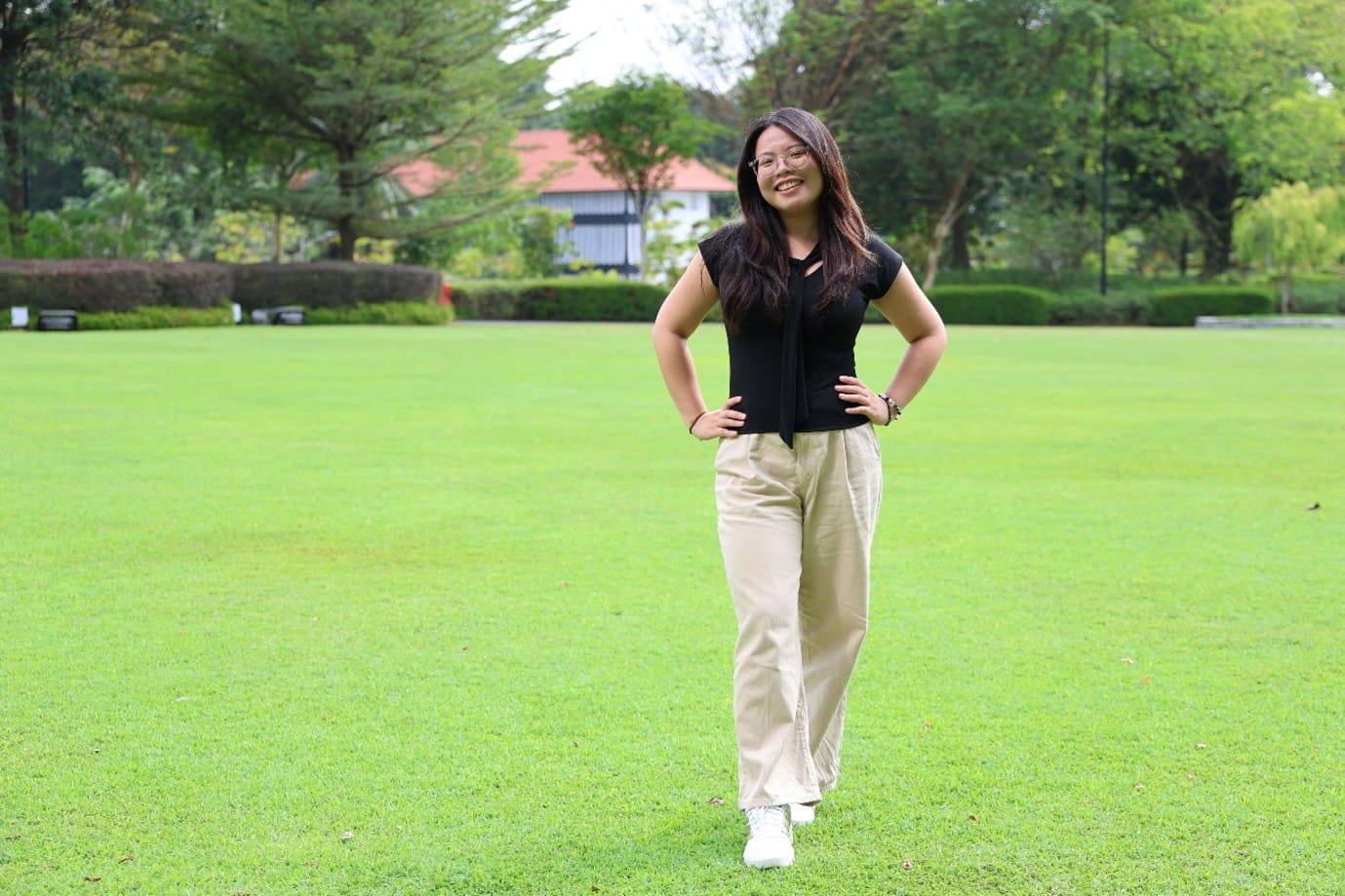 A smiling woman poses confidently with her hands on her hips in a lush, sunny park.